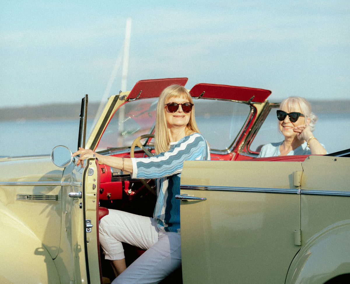 Two women in sunglasses seated in a cream vintage convertible by the water, one wearing a blue wave-stripe knit top