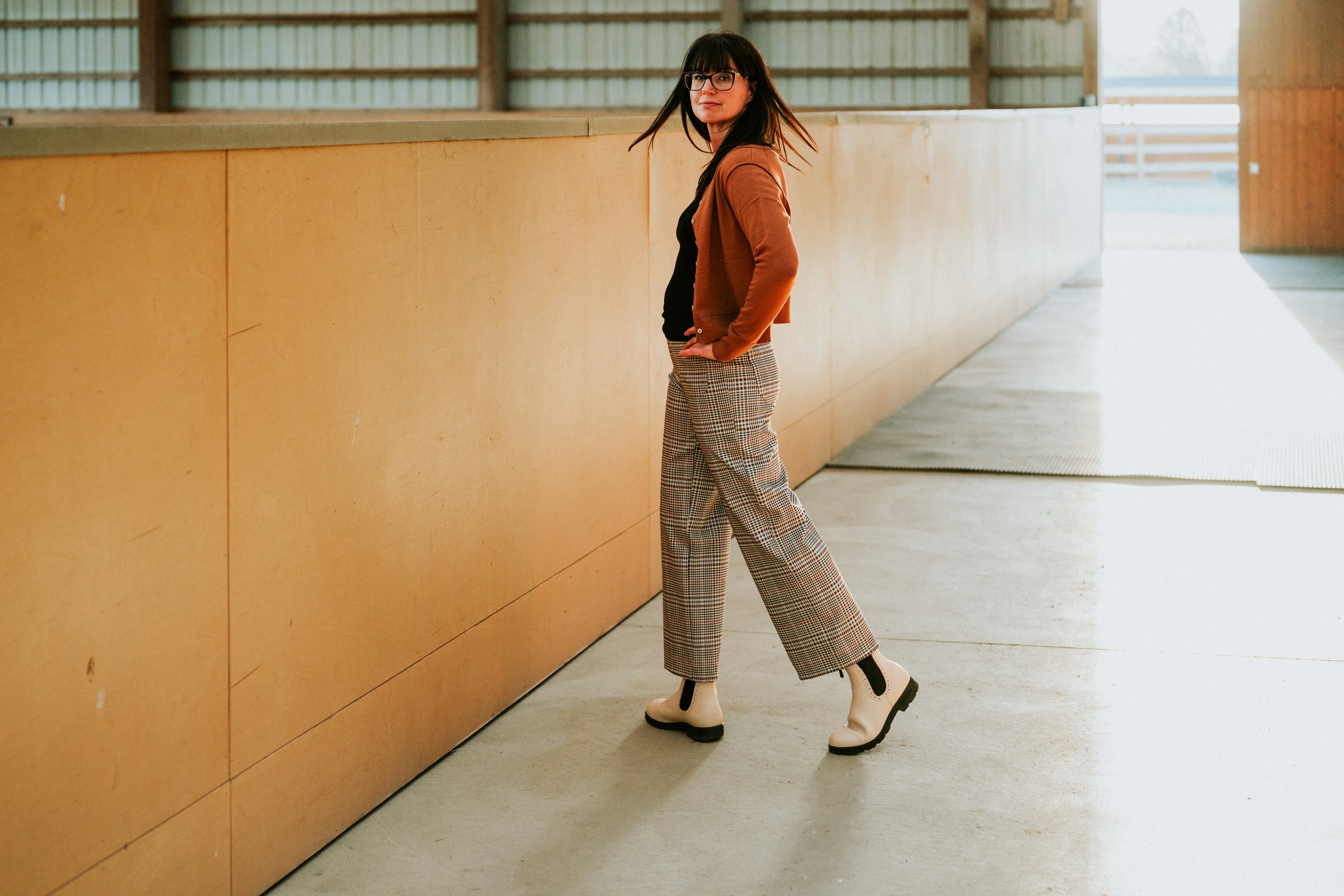 Woman in check wide-leg trousers and a rust cardigan walking through a covered riding arena
