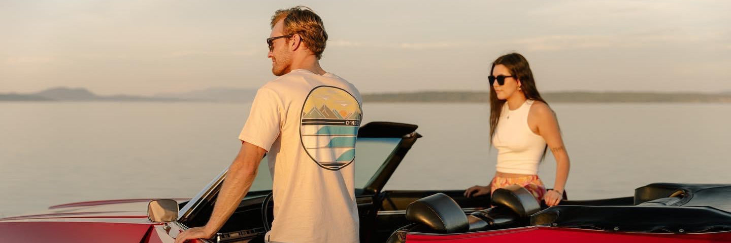 A man in a cream tee and jeans holds open the door of a red Impala convertible at golden hour, a woman seated behind him