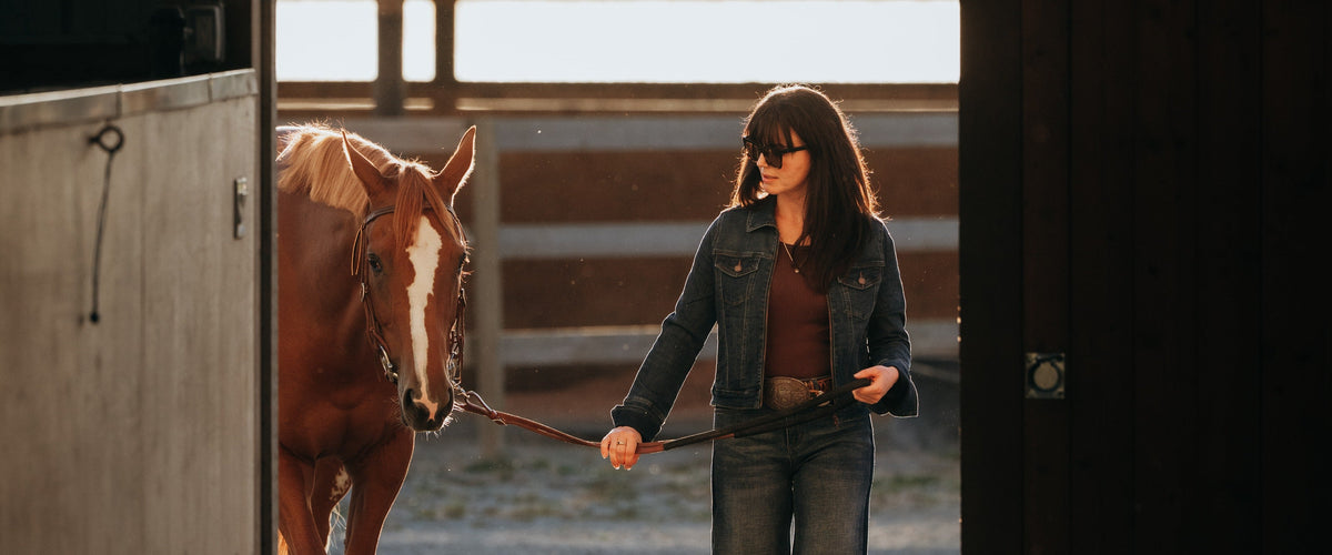 Woman in a denim jacket and jeans leading a chestnut horse through a sunlit barn doorway