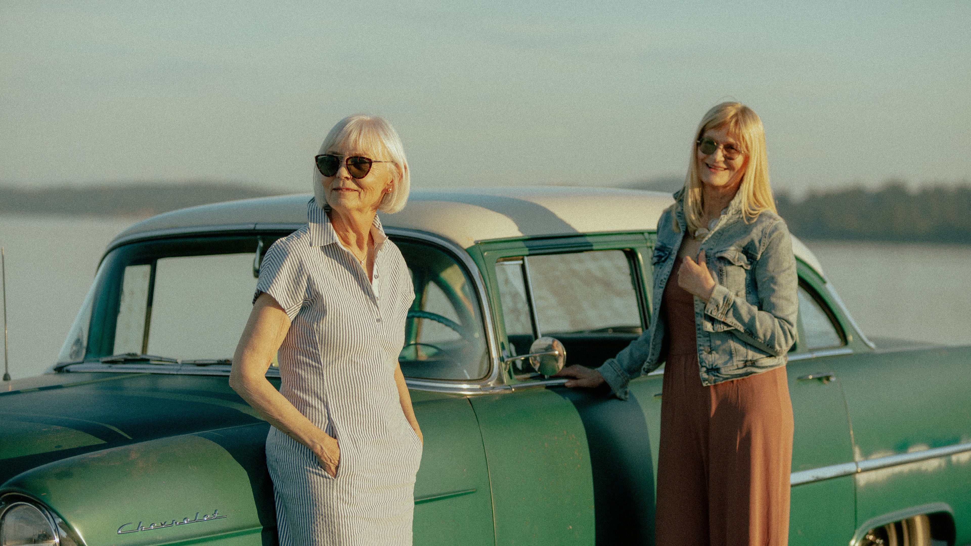 Two women in their sixties and seventies beside a vintage green Chevrolet at the water's edge in Sidney, British Columbia, at golden hour
