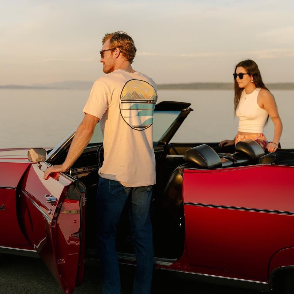 A man in a printed tee beside a red vintage Impala convertible at the water's edge at golden hour, with a woman seated in the car behind him