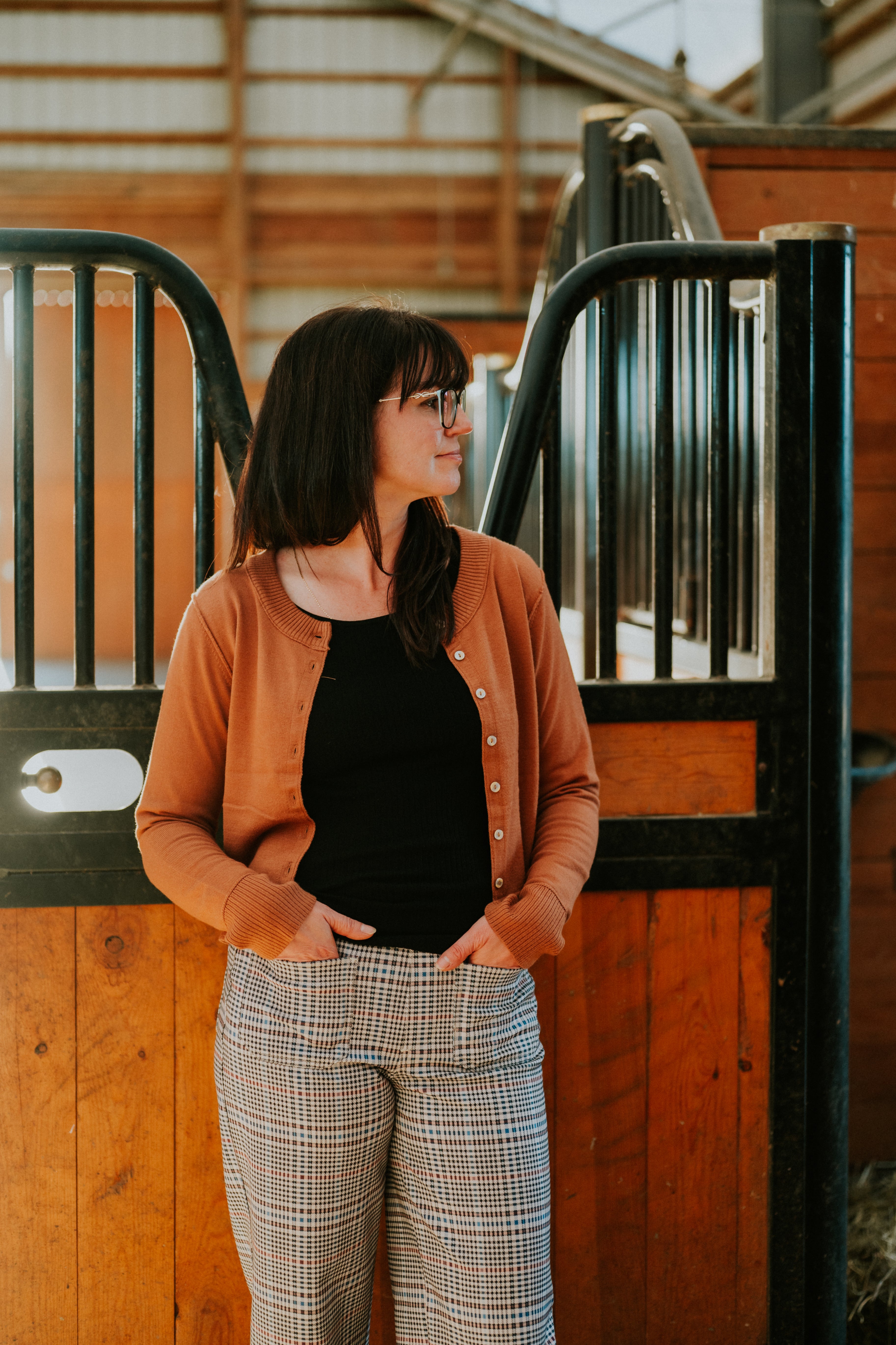 Woman in a rust button-front cardigan and check trousers standing by a stable door
