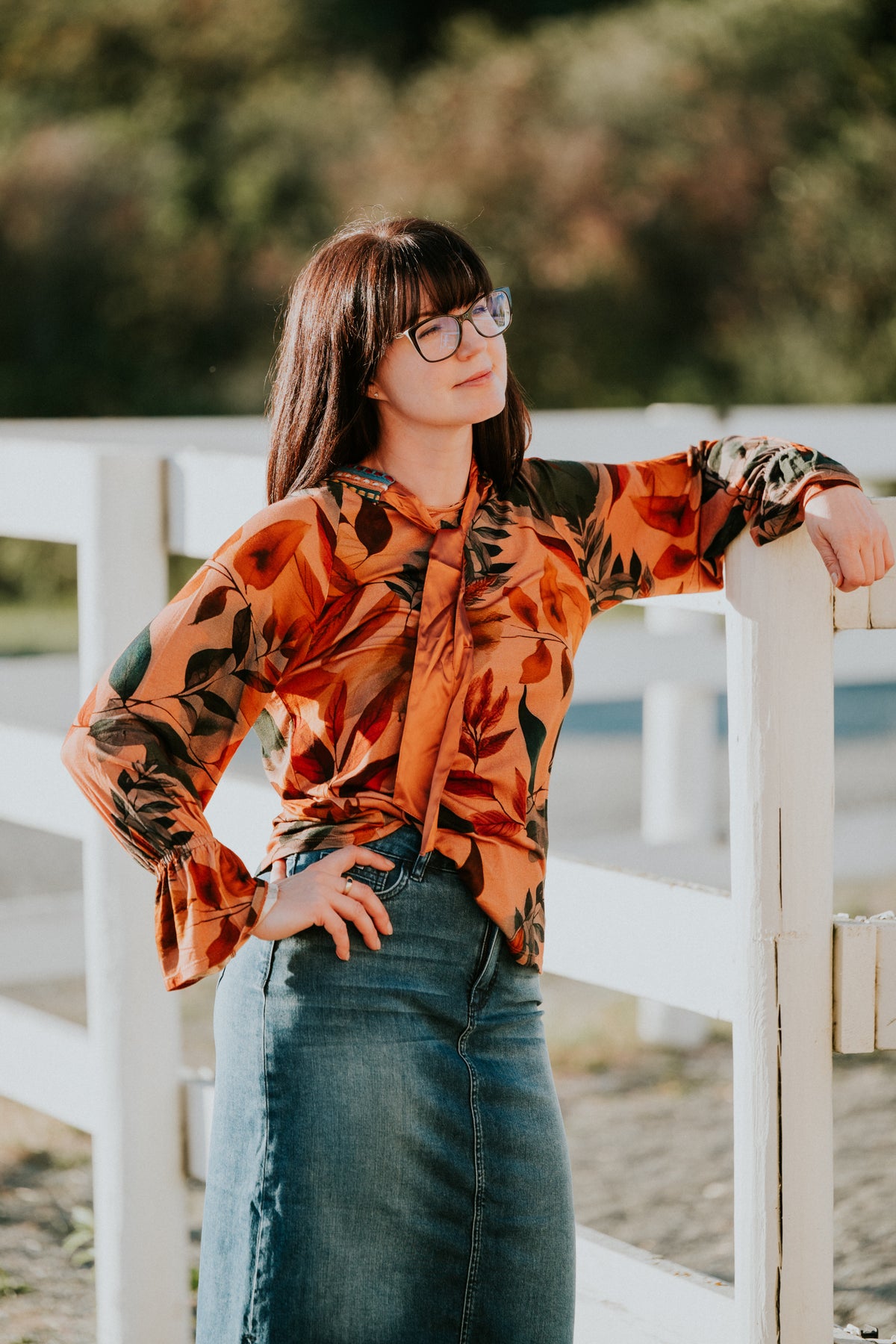 Woman in an autumn floral bell-sleeve blouse and denim skirt leaning on a white paddock fence