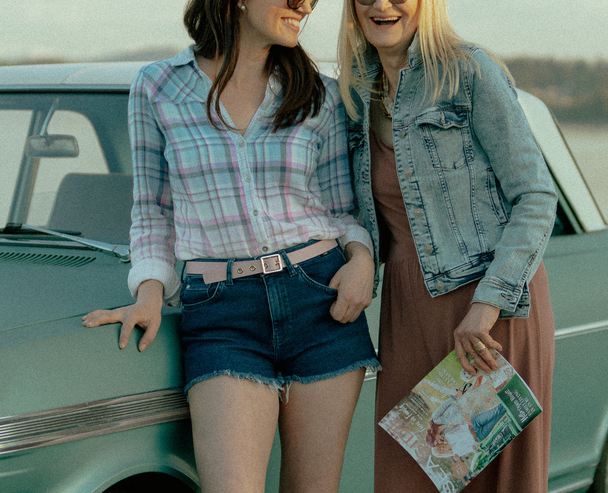 Two women laughing together beside a teal vintage Chevrolet, one in a plaid shirt and one in a denim jacket