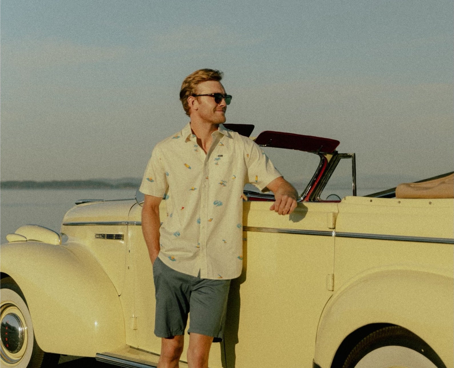 A man in a printed short sleeve shirt leaning against a cream vintage convertible by the water at golden hour