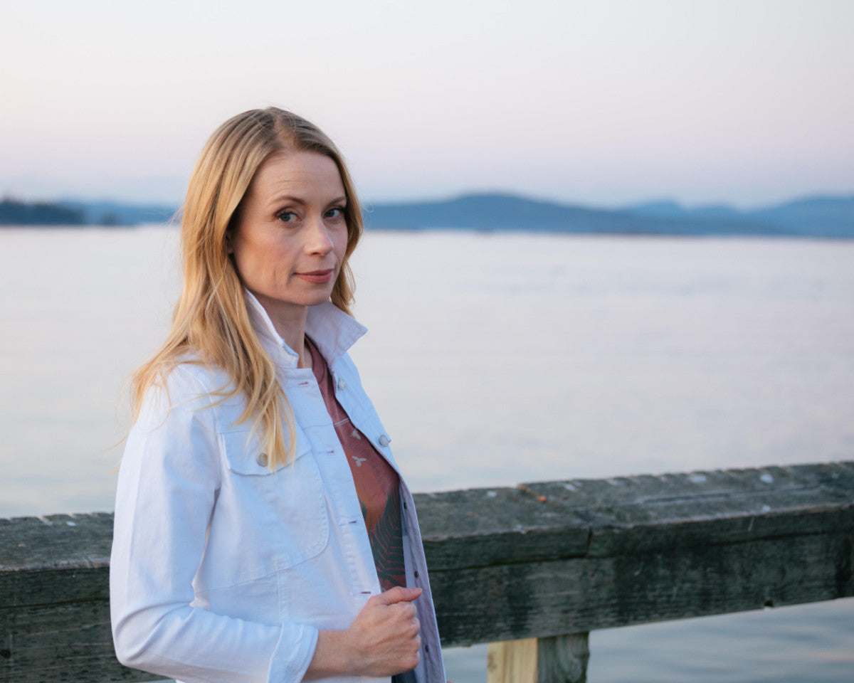 A woman in a white denim jacket on a wooden pier at dusk, with the water and hills behind her